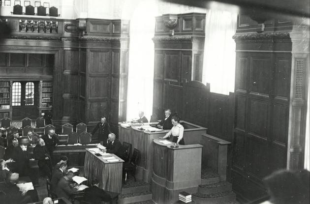 A woman is standing on a podium in a paneled room. Everyone else in the room are men.